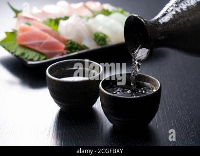 Sake wird in einen Sake-Becher gegossen, der auf schwarzem Hintergrund steht. Sashimi im Rücken. Stockfoto