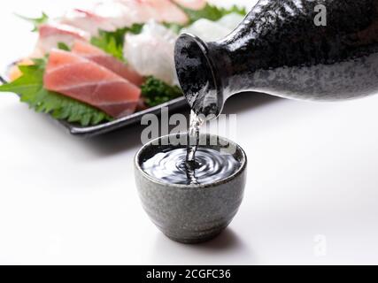 Der Sake wird in einen Sake-Becher gegossen, der auf weißem Hintergrund steht. Sashimi im Rücken. Stockfoto