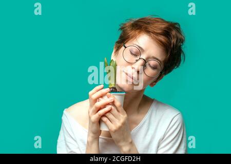 Kaukasische Frau mit Ingwer-Haar trägt Brille, Blick und lächeln auf die Kamera posiert mit Topfblume vor blauem Studio-Hintergrund. Stockfoto