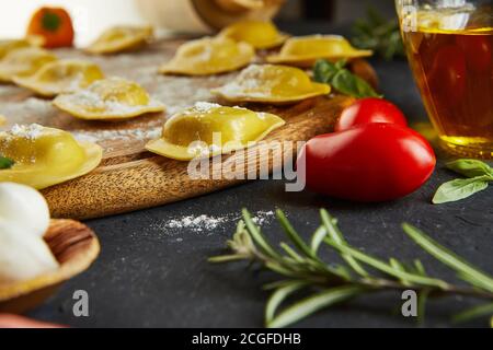Leckere Ravioli mit Mehl, Kirschtomaten, Sonnenblumenöl und Basilikum auf dunklem Grund. Der Prozess der Herstellung von italienischen Ravioli. Seitenansicht. Stockfoto