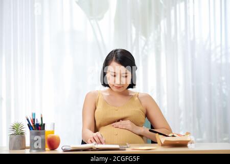 Schwangere junge Frau sitzt und mit digitalen Tablet auf dem Tisch Im Wohnzimmer und haben ein Frühstück und arbeiten Stockfoto