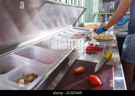 Ein Mann in einem Café bereitet Pizza zu. Die Zutaten für die Verpackung sind in Behältern verpackt und signiert. Stockfoto