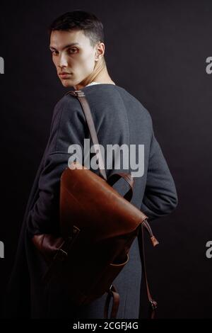 Lässige Outfits für Herren tragen blaue Jeans mit rotem kariertem Hemd, braunem Mantel und Ledertasche, die auf grauem Grunge-Hintergrund mit viel Platz und Lifestyle-Trave stehen Stockfoto