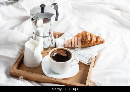 Holztablett mit Tasse schwarzen Kaffee, Kanne Milch und Croissant auf weißem Leinenbett. Frühstück im Bett mit Kaffee und Croissants. Stockfoto