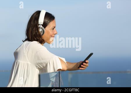 Erwachsene Frau mit Kopfhörer hört Musik auf einem Smartphone stehend Auf einem Balkon am Strand Stockfoto