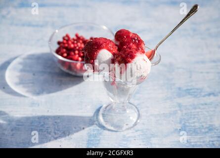 Eine Schüssel mit Eiskugeln und Marmelade aus roten Beeren auf blauem Hintergrund, Beeren in einer Glasvase, Sonnenschatten auf dem Tisch. Horizontale Ausrichtung Stockfoto