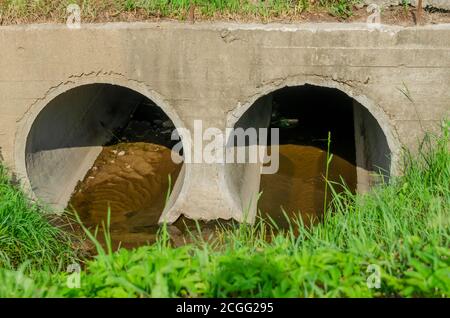 Zwei runde Rohre, durch die ein kleiner Fluss fließt. Diese Rohre laufen unter der Fahrbahn. Stockfoto