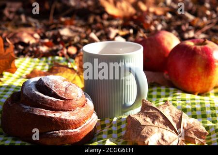 Stillleben im Herbst. Rote, reife Äpfel, eine Tasse Kaffee mit Milch in einer blauen Tasse, süßes Zimtbrötchen auf einer grün karierten Tischdecke vor einem Hintergrund Stockfoto