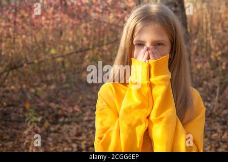 Schöne teen blonde Mädchen in einem leuchtend orange Sweatshirt bedeckt ihr Gesicht mit ihren Händen in einem Herbst Park. Herbstporträt im Wald gegen die b Stockfoto