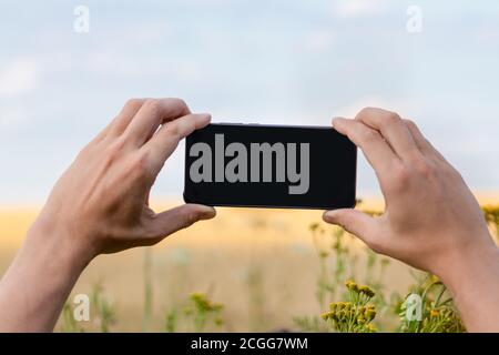 Halten Sie das Smartphone mit schwarzem Bildschirm mit zwei Händen horizontal In sonnigen Blumen Feld mit blauen Himmel natürliche Landschaft mockup Vorlage Stockfoto