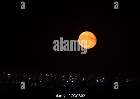 Großer, vollständig rot-oranger Mond, der am dunklen Nachthimmel über den Lichtern der Stadt aufgeht. Mystischer nächtlicher schwarzer Himmel mit großem glühenden Mond Stockfoto