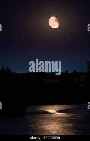 Mond scheint auf dunkelblauem Nachthimmel über still See Wasseroberfläche mit Reflexion und Stadt Silhouetten. Abnehmende Gibbous Phase. Langzeitbelichtung, vertikal Stockfoto