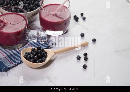 Zwei Gläser Vitamin-Drink Blaubeer Smoothie auf hellem Hintergrund, neben einer karierten Serviette, einem Holzlöffel mit Beeren, einem Teller ber Stockfoto