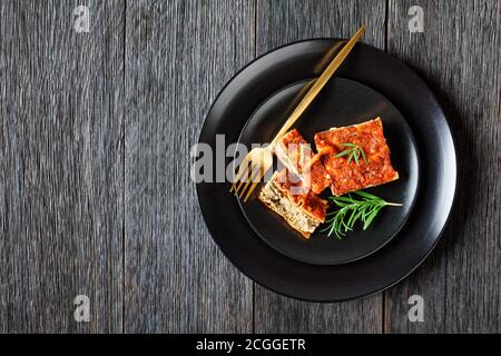 Stück Lasagne mit Champignon und Tomaten vegetarische Bolognese serviert auf einem schwarzen Teller mit Gabel und frischem Rosmarin auf einem dunklen rustikalen Holztisch, f Stockfoto