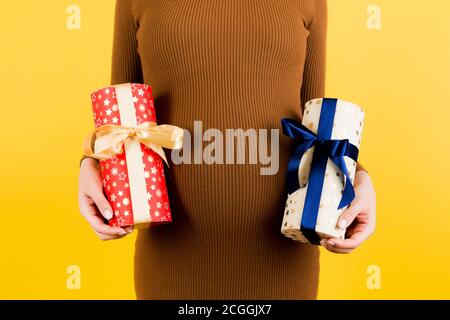 Zugeschnittenes Bild von schwangeren Frau in braunem Kleid mit zwei Geschenk-Boxen auf gelbem Hintergrund. Ist es ein Junge oder ein Mädchen? Warten auf Zwillinge. Schwangerschaft Promi Stockfoto