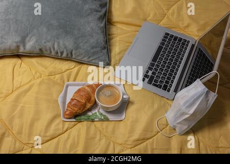 Layout, Stillleben, Laptop auf einer gelben Decke, Kaffee, Brötchen, medizinische Maske Corontin Konzept, Remote-Arbeit, Training während Coronavirus. Hochwertige Fotos Stockfoto