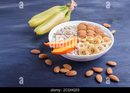 Ein Teller mit Müsli, Mandeln, Banane, Persimmon auf schwarzem Holzgrund. Nahaufnahme. Platz kopieren Stockfoto