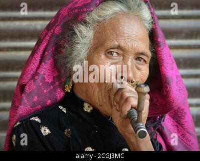 Elegante ältere Nepali Magar Frau mit traditionellem Nasenschmuck raucht eine Pfeife mit lokalem Tabak. Stockfoto