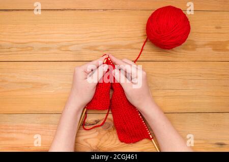 Damenhänden mit Stricknadeln und einer roten Garnkugel auf einem Holzhintergrund. Stockfoto