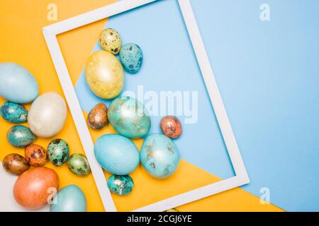 Bemalte Ostern große und kleine Eier auf einem blauen und gelben Hintergrund, weißen Holzrahmen daneben. Freier Speicherplatz für Text. Frohe Ostern! Stockfoto