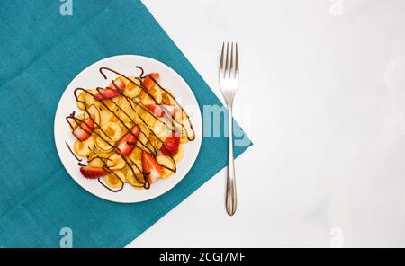 Leckeres und gesundes modisches Frühstück für die ganze Familie. Mini-Pfannkuchen auf einem weißen Teller mit Bananenscheiben, Erdbeeren und Schokoladensauce. Stockfoto