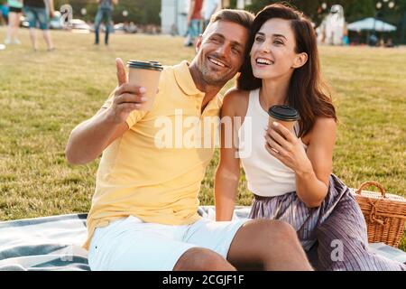 Portrait von aufgeregt im mittleren Alter Paar Mann und Frau trinkt Kaffee zum Mitnehmen und Zeigefinger beim Sitzen auf Gras im Park Stockfoto