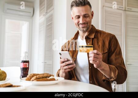 Bild von schönen fröhlichen Mann mit Kreditkarte und mit Smartphone, während Sie in der weißen Küche zu Mittag essen Stockfoto