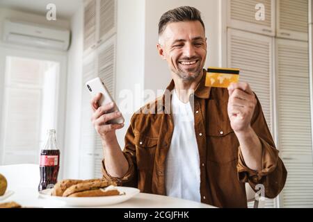 Bild von schönen fröhlichen Mann mit Kreditkarte und mit Smartphone, während Sie in der weißen Küche zu Mittag essen Stockfoto