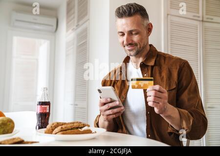 Bild von schönen fröhlichen Mann mit Kreditkarte und mit Smartphone, während Sie in der weißen Küche zu Mittag essen Stockfoto