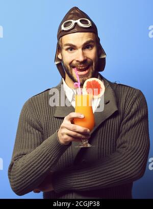 Schöner bärtiger Flieger Mann mit langem Bart auf lächelndem Gesicht Hält Glas alkoholischen Cocktail in grau gestrickt Pullover mit hut und Brille auf blauem Studiohintergrund Stockfoto
