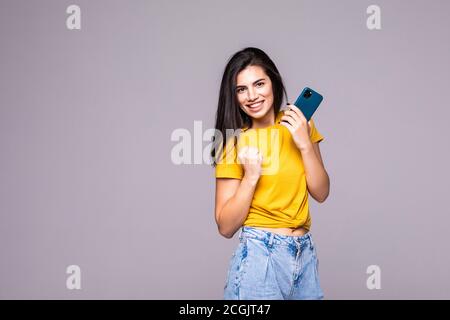 Glückliche junge Frau, die ihre Fäuste in Gewinnergeste zusammenhält und die Kamera auf grauem Hintergrund anschaut Stockfoto