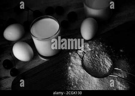 Schwarz-Weiß-Foto von Mehl in Metallsieb, Eier. Milch und Beeren Stockfoto