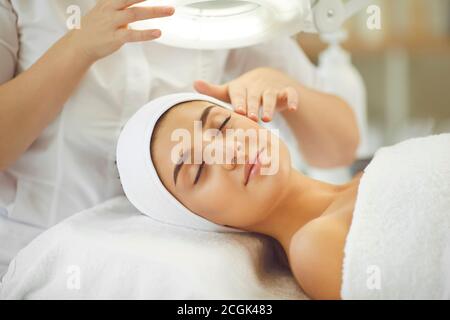 Lächelnde junge Womans Gesicht unter Licht bekommen und genießen Gesichtsbehandlung Massage vom Kosmetologen Stockfoto