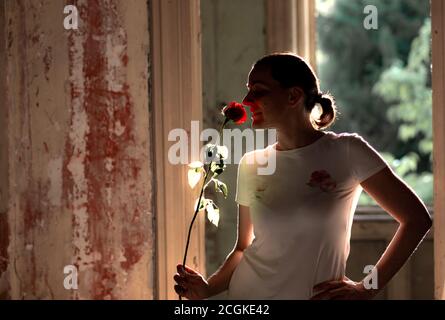 Silhouette einer jungen Frau, die eine rote Rose riecht, in einem ruinierten, verlassenen Haus. Glück. Schlechte Zeiten - gute Zeiten Konzept. Hintergrundbeleuchtung. Stockfoto