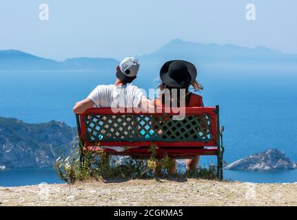 Ein junges Paar, das Hüte trägt und von einer Bank auf der griechischen Insel korfu in griechenland aus über das Meer blickt. Stockfoto