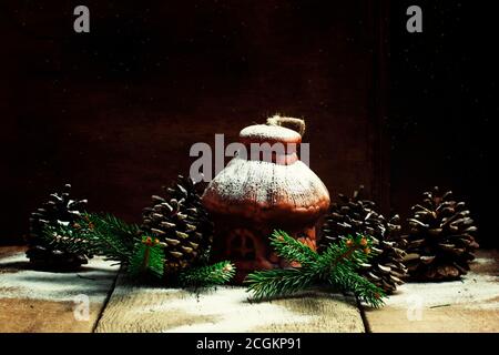 Weihnachtshaus aus Ton mit Tannenzapfen und Tannenzweigen unter dem Schnee, Vintage-Holzhintergrund, selektiver Fokus Stockfoto