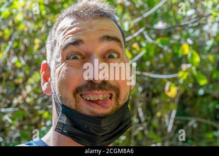 Porträt eines Mannes mittleren Alters mit der Maske, um sich vor dem Coronavirus zu schützen gesenkt, um im Wald zu lächeln. Naturkonzept Stockfoto