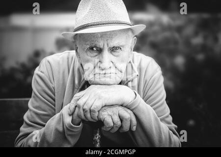 Trauriger, wütender alter Mann im Hut sitzt in einem Freiluftgarten. Das Konzept von Einsamkeit und einsamem Alter. Schwarz-Weiß-Porträt Stockfoto