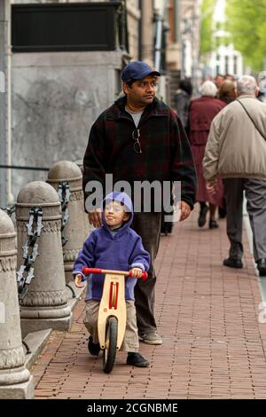 Amsterdam, Niederlande 05/15/2010: Ein mittelöstlicher oder indischer Vater ist auf seinen Sohn, der auf dem Bürgersteig auf einem belebten U-Rad fährt, aufgesetzt Stockfoto