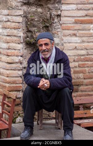 Adana, Türkei 12/26/2009: Porträt eines älteren türkischen Mannes in Salwar und einer Jacke, der auf einem Holzstuhl sitzt, während er lächelt und eine Cig raucht Stockfoto