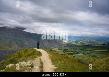 Ein schwarz getragener Mann steht auf einem Hügel, um im Sommer in Otago, Neuseeland, einen Panoramablick auf den Arrow Junction Lookout Point zu genießen. Stockfoto