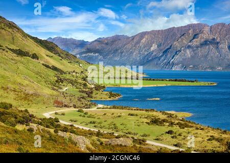 Blick auf die Berge und den See Hawea. Im Sommer gibt es grünes Gras und blauen Himmel mit schönen Wolken am Hals, Otago, Neuseeland. Stockfoto