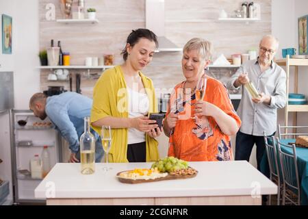 Mutter und Tochter benutzen Smartphone in der Küche. Ältere Frau mit einem Glas Wein. Alter Mann, der die Flasche anschaut. Stockfoto