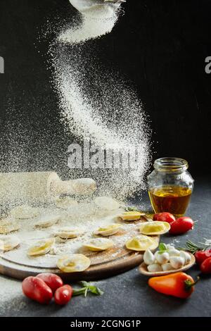 Das Mehl fällt auf die Ravioli. Köstliche rohe Ravioli mit Käse und Mehl, Kirschtomaten, Sonnenblumenöl und Basilikum auf dunklem Grund. Stockfoto