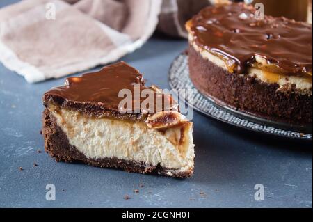 Ein Stück Käsekuchen Snickers mit Schokolade, Karamell, Erdnusspaste, Nougat und Erdnusskuchen auf blauem Beton-Tischhintergrund. Stockfoto