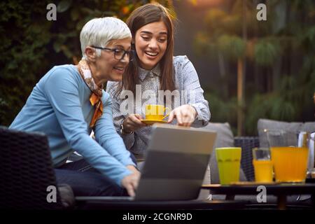 Alte fröhliche Mutter und junge Erwachsene Tochter, die Inhalte auf dem Laptop anschauen und Kaffee trinken Stockfoto