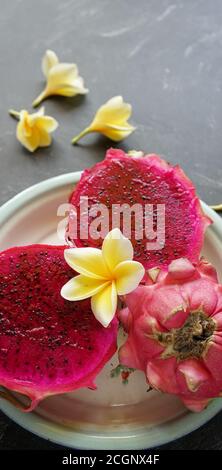 Zwei Scheiben rote Drachenfrucht und eine ganze Drachenfrucht, serviert auf einem Tablett und verziert mit Frangipani Blumen, auf einem Steinblock Stockfoto