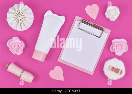 Flaches Lay mit leerem Clipboard und verschiedenen niedlichen Gegenständen wie Rosenblumen, cremefarbenem Rohr in Form von Katze, Papierherzen und Puderpuff auf rosa Hintergrund Stockfoto