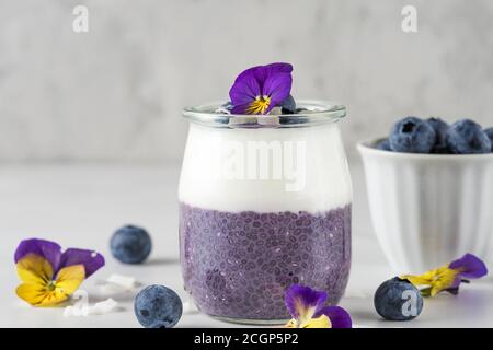 Gesundes Frühstück mit Heidelbeer-Parfaits mit frischem Obst, Kokosjoghurt, Chia-Pudding und essbaren Blumen auf weißem Tisch Stockfoto