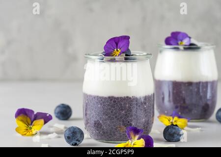 Heidelbeer Joghurt Parfait mit Chia Pudding, Kokosjoghurt, frische Beeren und Blumen in Gläsern für gesunde Ernährung Frühstück auf weißem Marmortisch mit Stockfoto
